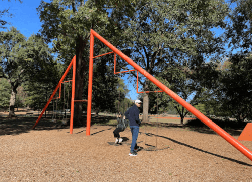 A child swings on a playground while an adult stands nearby, surrounded by trees and a clear blue sky.