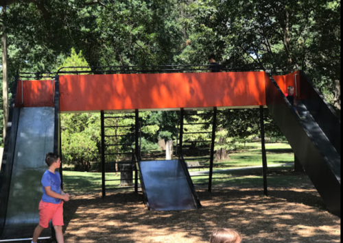 A playground structure with two slides and climbing features, surrounded by trees and grass. Children are playing nearby.