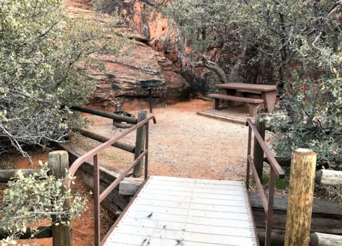 A wooden bridge leads to a picnic area with a table and grill, surrounded by rocky terrain and shrubs.