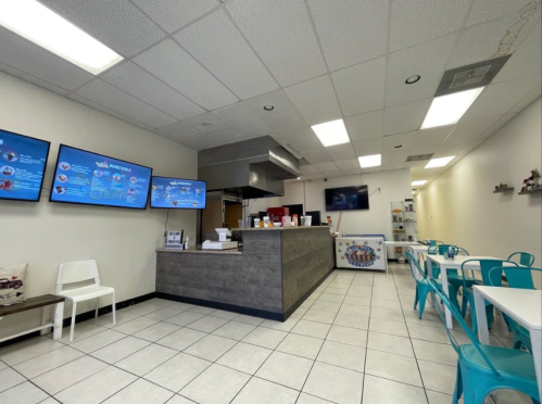 Interior of a casual eatery featuring a service counter, digital menus, and blue chairs at white tables.