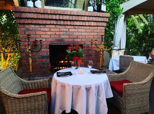 Cozy outdoor dining setup with a table, candles, and a brick fireplace surrounded by greenery and elegant decor.