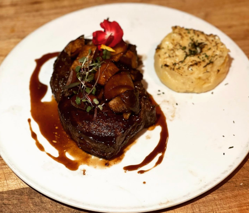 A plated dish featuring a steak topped with mushrooms and sauce, accompanied by a creamy side and a flower garnish.