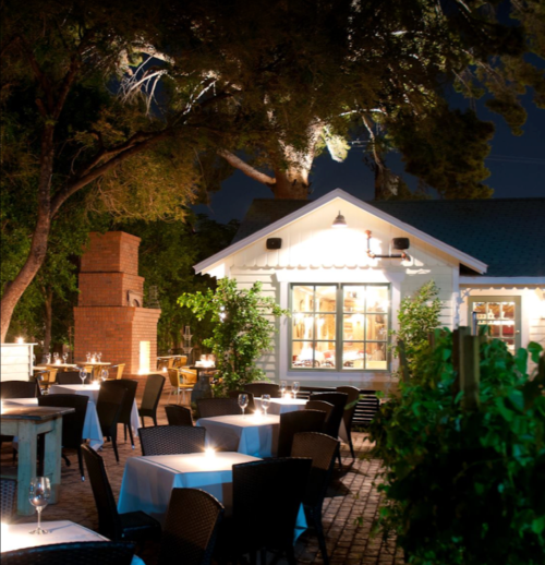 A cozy outdoor restaurant at night, featuring tables with white tablecloths and a charming lit-up building.