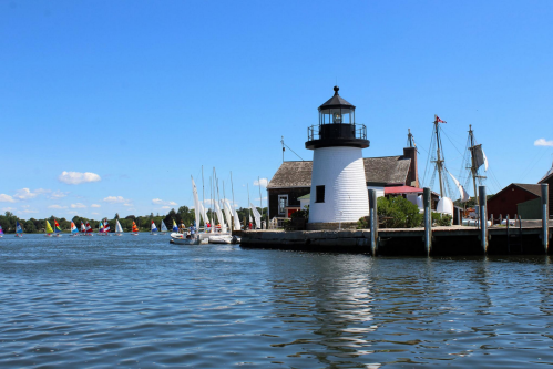 A scenic waterfront view featuring a lighthouse, sailboats, and clear blue skies.