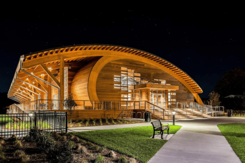 A modern wooden building with a curved roof, illuminated at night, surrounded by green grass and a pathway.