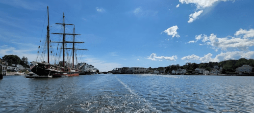 A scenic view of a harbor with a tall ship, calm water, and a clear blue sky dotted with clouds.