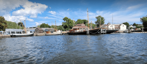 A scenic view of boats docked along a river, with trees and buildings in the background under a blue sky.