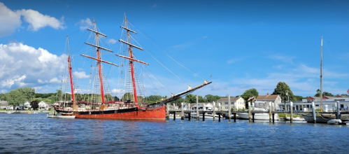 A tall ship with red sails docked by a waterfront, surrounded by buildings and a clear blue sky.