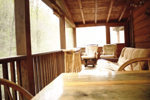 Cozy porch with wooden furniture, including chairs and a sofa, surrounded by trees and a misty atmosphere.