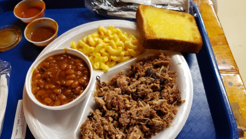 A tray with pulled pork, macaroni and cheese, baked beans, and a slice of buttered bread.