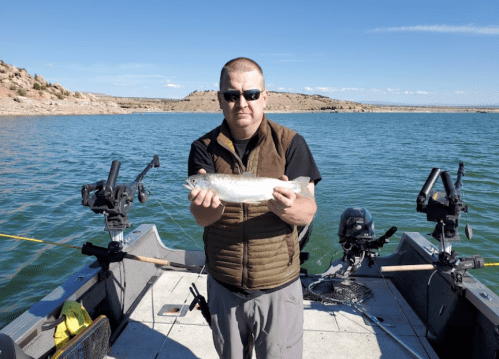 A person holding a fish on a boat in a lake, with mountains and clear blue sky in the background.