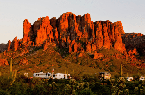Sunset over rugged red rock formations with RVs parked in a desert landscape. Cacti dot the foreground.