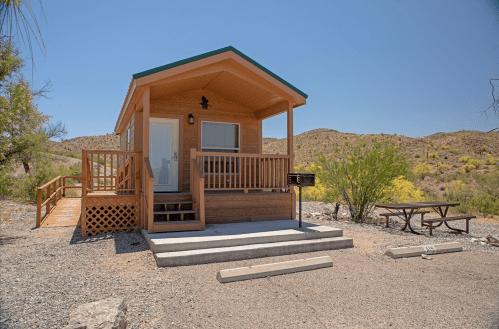 A small wooden cabin with a porch, surrounded by desert landscape and a picnic table nearby.