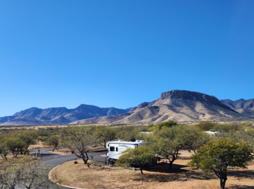 A scenic view of mountains under a clear blue sky, with an RV parked among trees in the foreground.