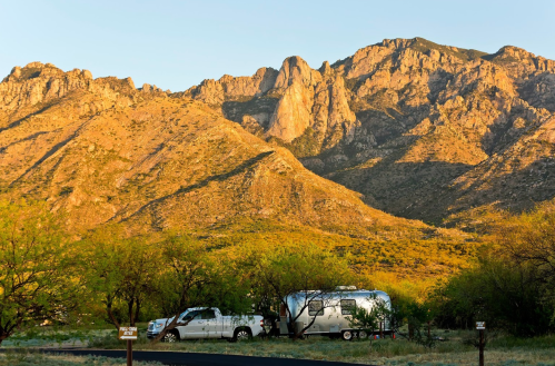 A silver Airstream trailer parked near a mountain range at sunset, surrounded by green trees and a clear sky.