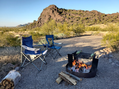 Two camping chairs beside a fire pit with a fire, surrounded by desert landscape and mountains in the background.