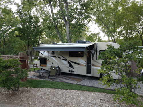 A parked RV with an awning, surrounded by trees and a small outdoor seating area in a campground.
