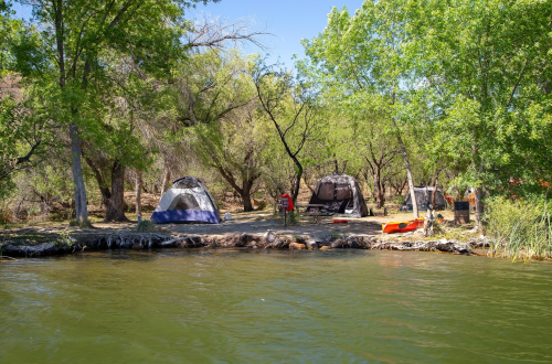 Tents set up by a riverbank, surrounded by trees, with a kayak and a campfire area visible nearby.