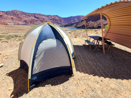 A tent set up near a river, with rocky cliffs in the background and a shaded picnic area nearby.