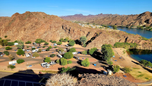 Aerial view of a campground near a river, surrounded by rocky hills and mountains under a clear blue sky.