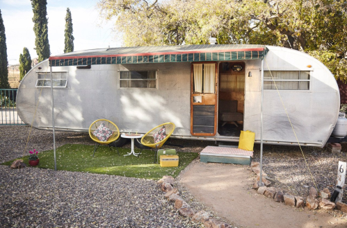 A vintage trailer with a green awning, surrounded by gravel and plants, featuring colorful chairs and a welcoming entrance.