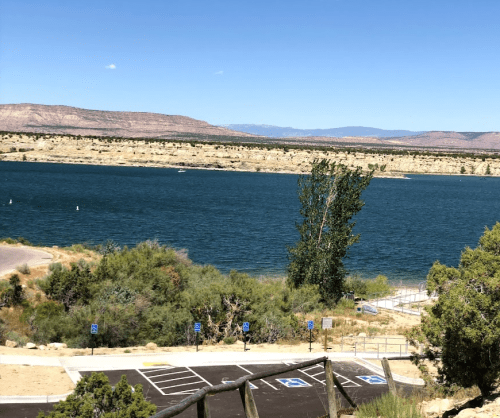 A serene lake surrounded by mountains, with a parking area and trees in the foreground under a clear blue sky.