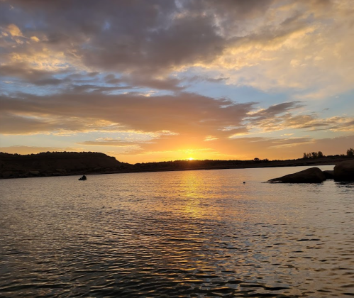 A serene sunset over a calm river, with clouds reflecting warm colors and rocky shores in the foreground.