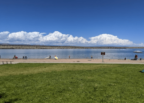 A serene beach scene with people enjoying the water, green grass in the foreground, and fluffy clouds in a blue sky.
