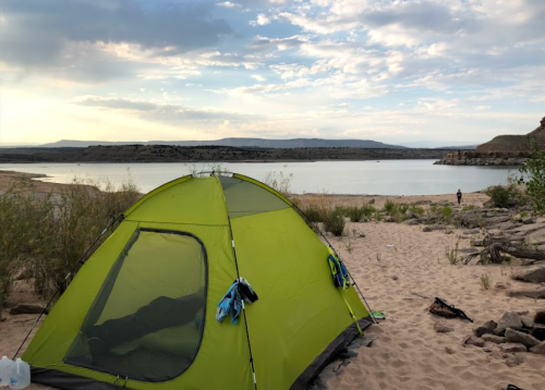 A green tent set up on sandy ground by a lake, with mountains in the background and a cloudy sky.