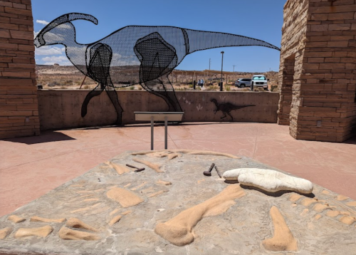 A wireframe dinosaur sculpture stands behind a fossil display in a desert landscape.