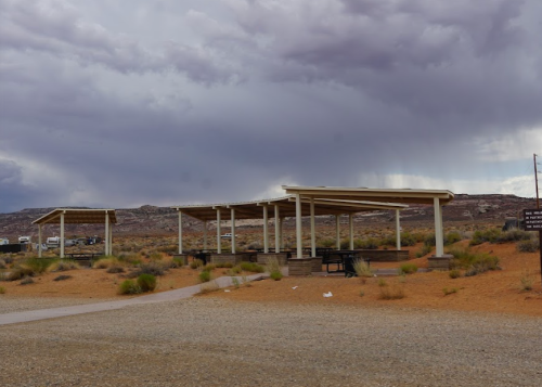 Sheltered picnic area in a desert landscape under a cloudy sky, with sparse vegetation and gravel paths.