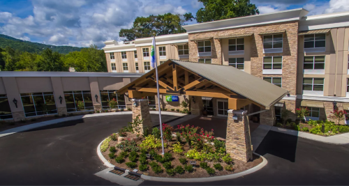 A modern hotel entrance with a wooden canopy, surrounded by landscaped gardens and mountains in the background.