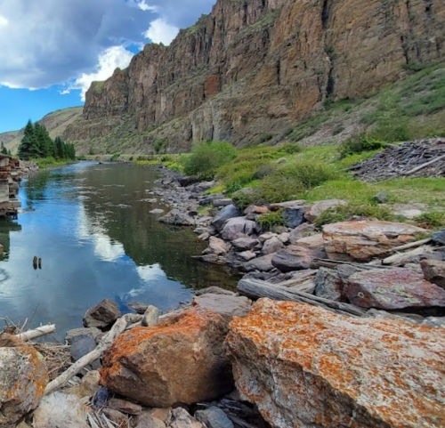 A serene river flows between rocky banks, surrounded by steep cliffs and lush greenery under a partly cloudy sky.