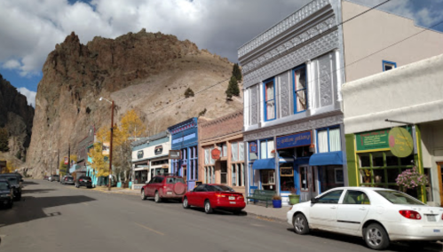 A quaint street lined with colorful shops and cars, set against a backdrop of rocky mountains under a cloudy sky.