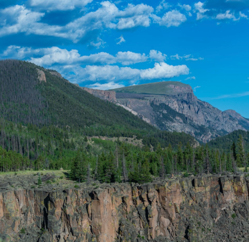 A scenic view of rugged mountains and cliffs under a blue sky with scattered clouds, surrounded by lush green forests.