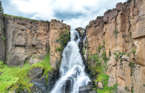 A stunning waterfall cascades down rocky cliffs, surrounded by lush greenery and a cloudy sky. Two people stand at the top.