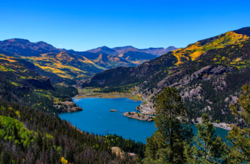 A scenic view of a lake surrounded by mountains and colorful autumn foliage under a clear blue sky.