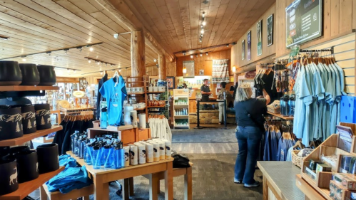 A cozy gift shop with wooden decor, featuring clothing, souvenirs, and a customer browsing the merchandise.