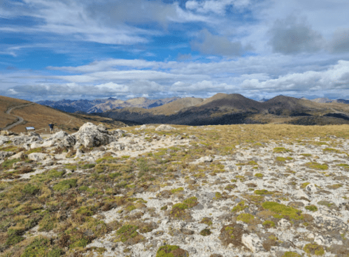 A panoramic view of rolling hills and mountains under a partly cloudy sky, with rocky terrain in the foreground.
