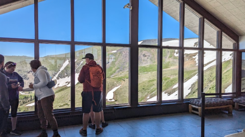 A group of people stands by large windows, overlooking a scenic mountain view with snow and greenery.