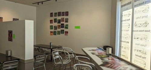 A bright gallery space with artwork on the walls, tables, and chairs, featuring a window display of printed materials.