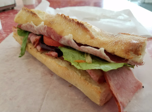 A close-up of a sandwich with layers of ham, lettuce, and tomato in a toasted baguette on a paper surface.