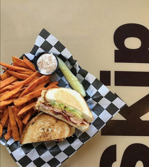 A sandwich with lettuce and meat, served with sweet potato fries, a pickle, and a small cup of dipping sauce on a checkered tray.