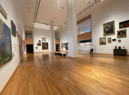 A spacious art gallery with wooden floors, featuring various paintings and sculptures, and a visitor observing the exhibits.