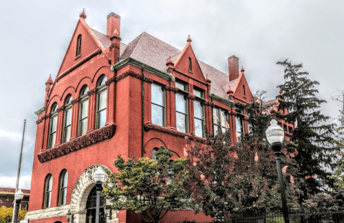 Historic red brick building with ornate architectural details and large windows, surrounded by trees and lampposts.