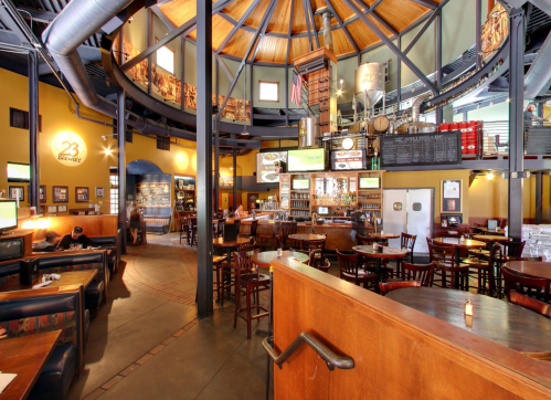 Interior of a spacious brewery with high ceilings, wooden tables, and a bar area, featuring brewing equipment in the background.