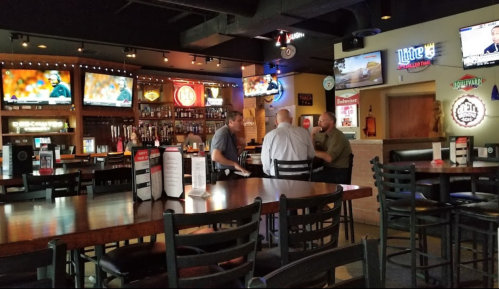 A cozy bar interior with patrons chatting, multiple TVs showing sports, and a well-stocked bar in the background.