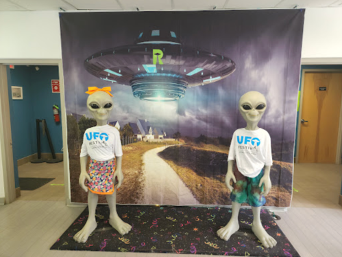 Two alien figures in T-shirts stand in front of a UFO-themed backdrop featuring a spaceship and a path.