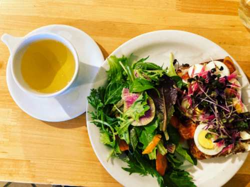 A plate with a colorful salad, a slice of toast topped with eggs and microgreens, and a cup of light-colored tea.
