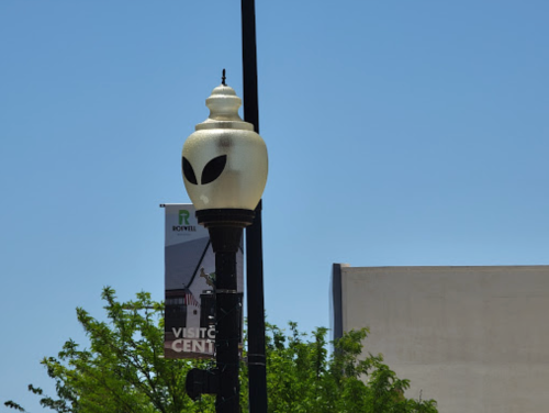 A street lamp with an alien-themed design against a clear blue sky, with a banner for a visitor center nearby.